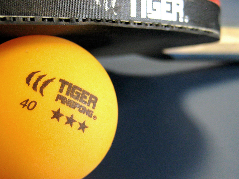 Orange ping pong ball on a table with a ping pong paddle on top of the ball.