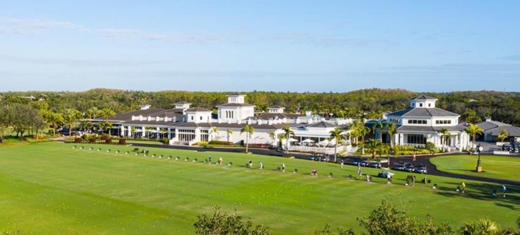 The image depicts a championship golf course with lush green fairways and strategically placed water hazards, while luxury homes are visible in the background, showcasing the upscale lifestyle of the Shadow Wood community in Bonita Springs, Florida. The scene reflects a tranquil setting perfect for golf enthusiasts and those seeking luxury living in a prime neighborhood.