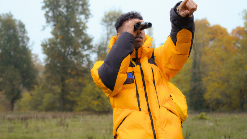 A person wearing a bright yellow puffer gorpcore jacket with black accents looks through binoculars and points into the distance, with trees and a field in the background.
