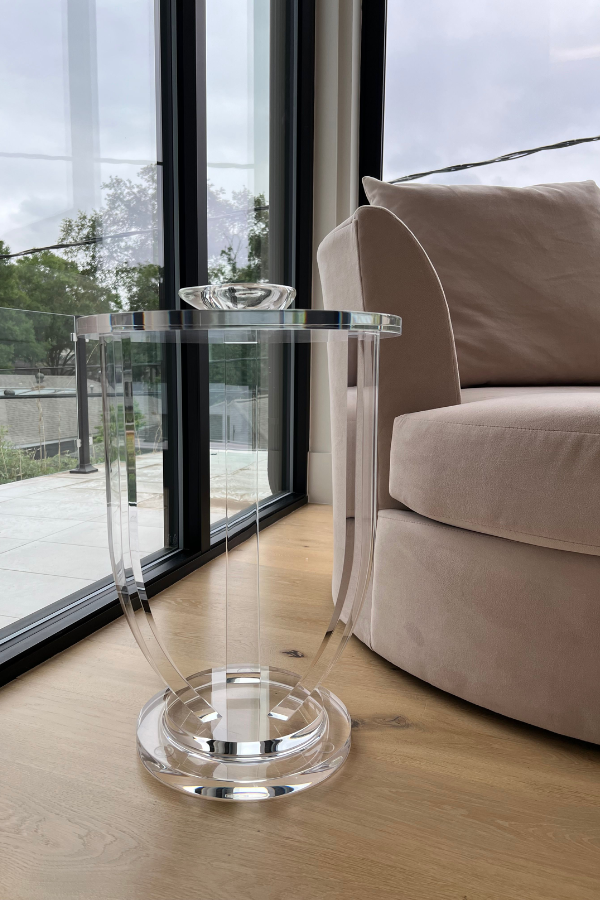 A clear acrylic side table beside a curved beige lounge chair, set against floor-to-ceiling windows overlooking an outdoor terrace.