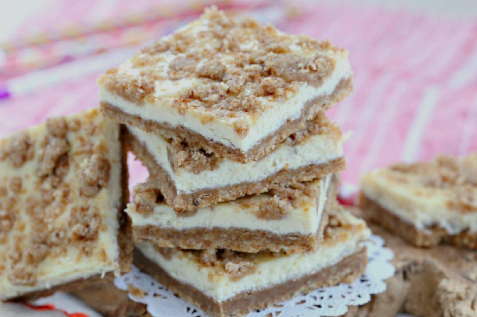 A stack of crumb-topped cheesecake bars with a graham crust, on a lace doily. The bars convey a homemade and inviting look, set against a pink-striped cloth.