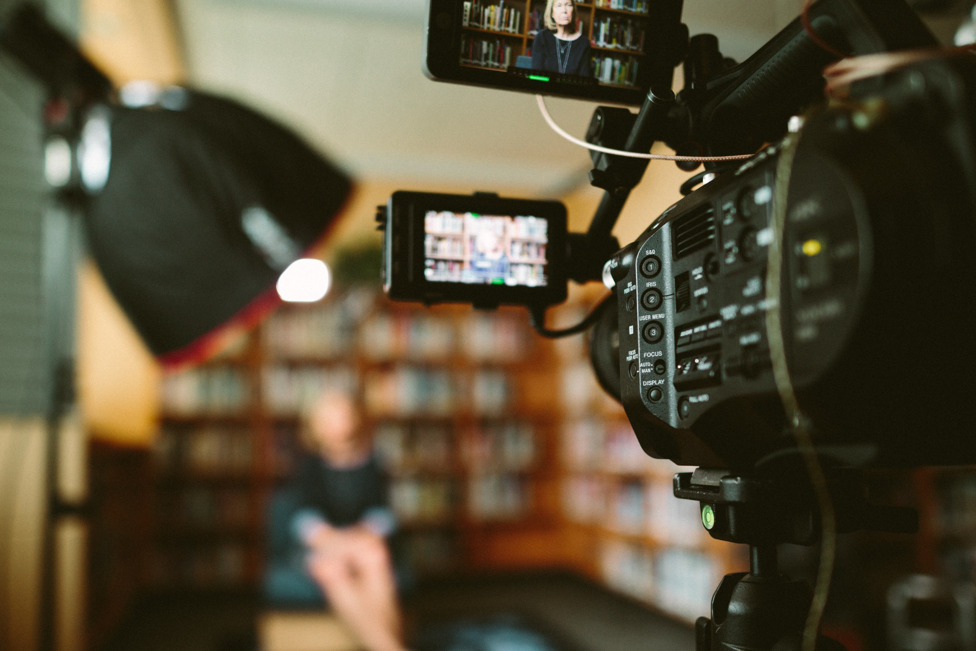 A professional video camera with dual monitors filming a person seated in front of a bookshelf in a softbox-lit studio setting.