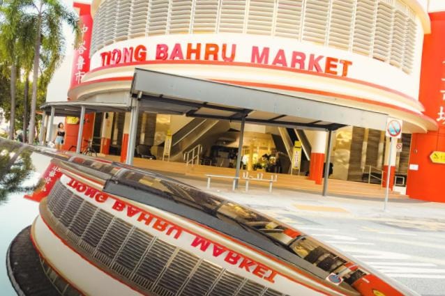 This image features the entrance to the Tiong Bahru Market in Singapore, highlighted by its distinctive curved architecture and bright red signage. The shot captures a unique perspective through a reflection on a nearby surface, mirroring the building's facade and the surrounding street scene.