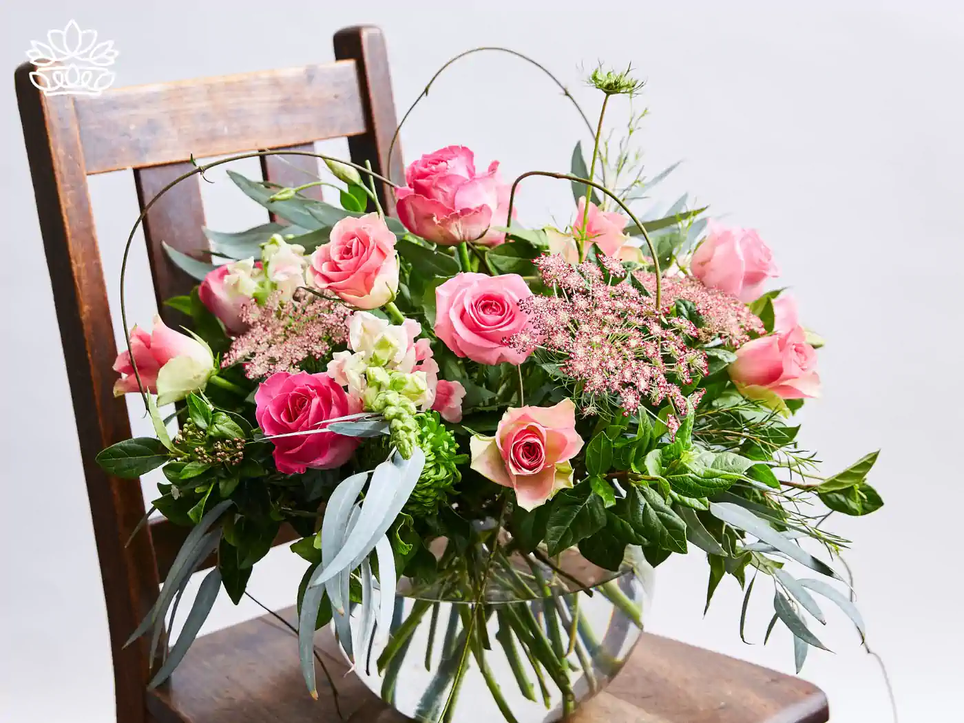 Artistic floral arrangement displayed on an antique wooden chair, featuring pink roses, blush dahlias, cascading green amaranthus, and whimsical curly willow branches. 