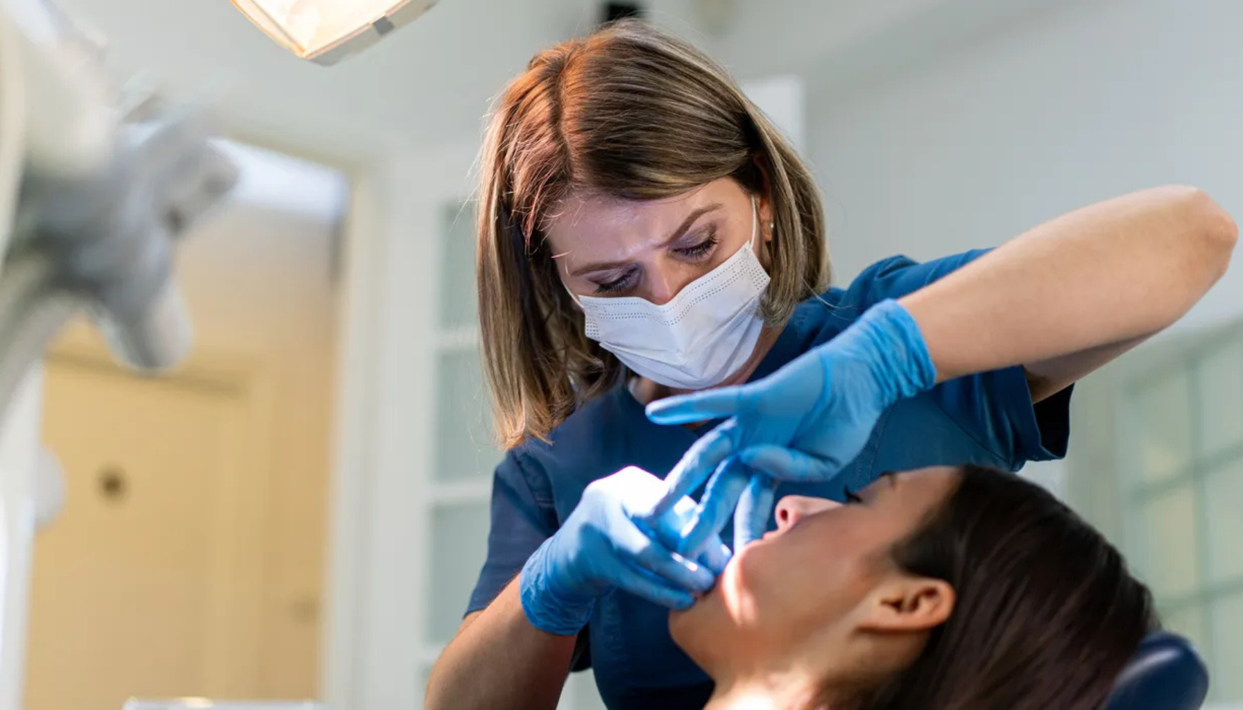 Tannlege med maske og blå hansker utfører tannundersøkelse på pasient som sitter i tannlegestolen med åpen munn.