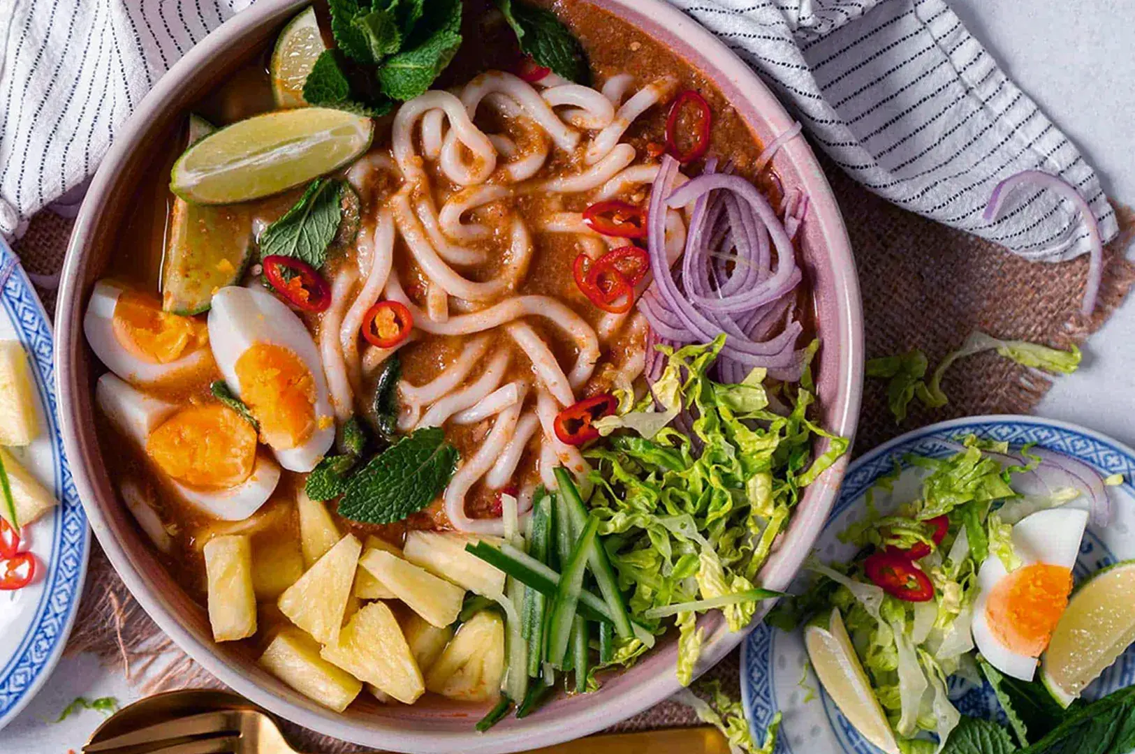 A vibrant bowl of noodle soup with sliced boiled eggs, pineapple, cucumber, mint, red onion, chilies, and lime wedges. A striped cloth adds a cozy touch.