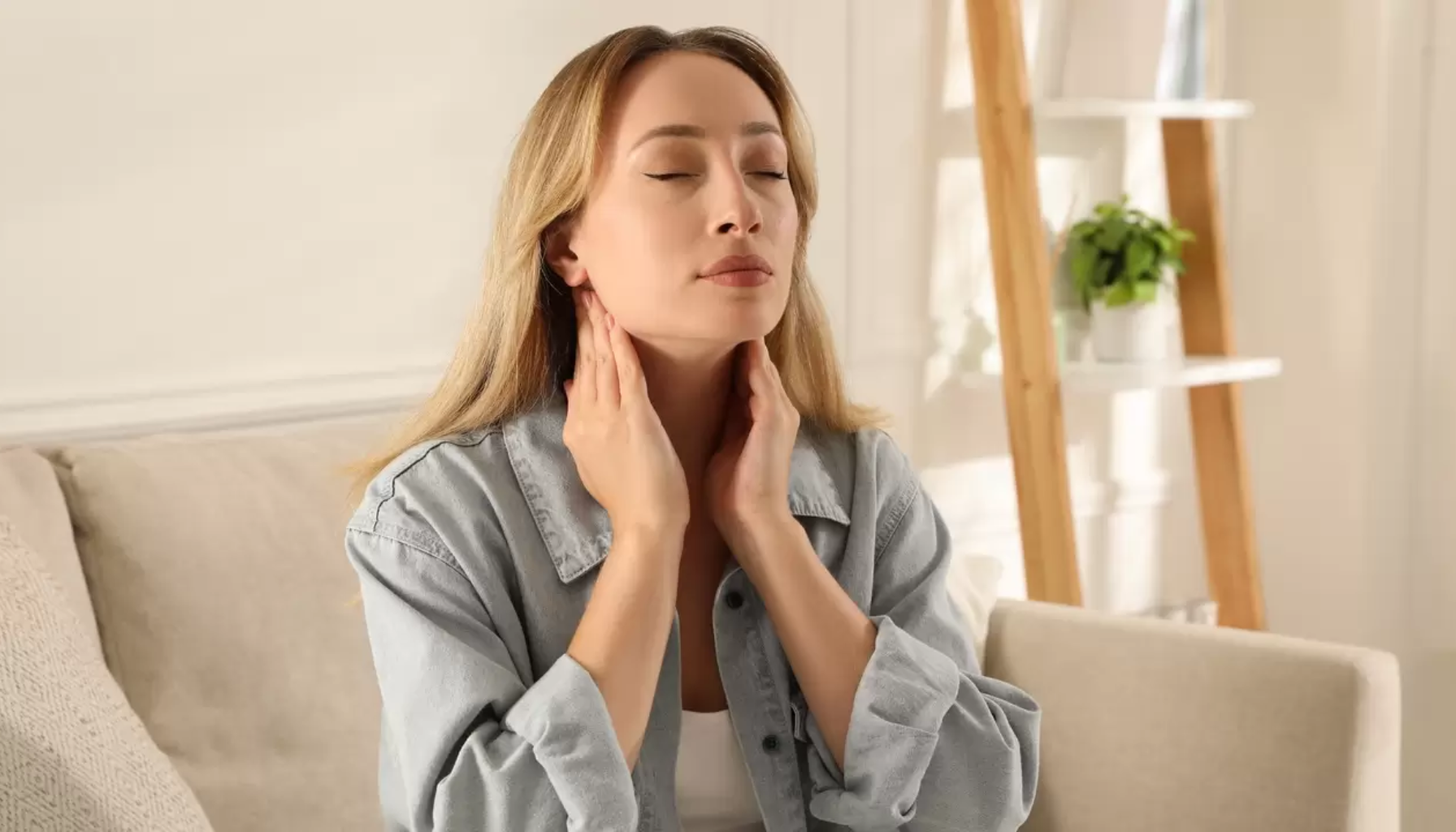 Kvinne som sitter hjemme og holder hendene på halsen, ser ut til å ha vondt eller ubehag i halsområdet.»