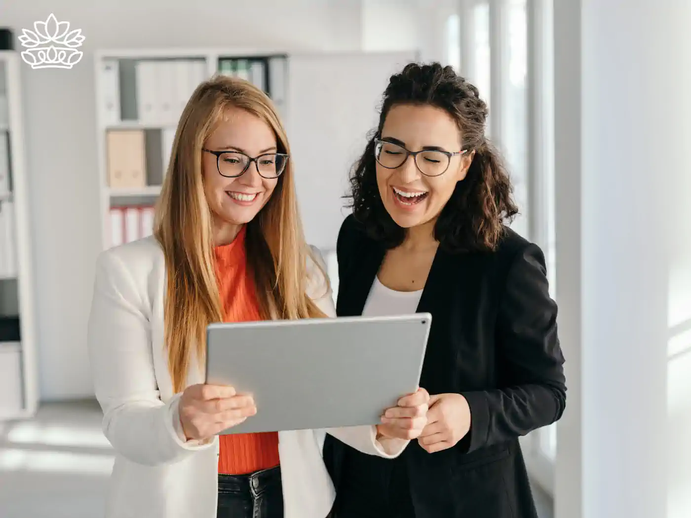 Two professional women in a bright office setting, smiling joyfully as they look at a tablet together, sharing a positive work-related experience. They are stylishly dressed, embodying a sense of partnership and success. Gift boxes for colleagues, delivered with heart by Fabulous Flowers and Gifts