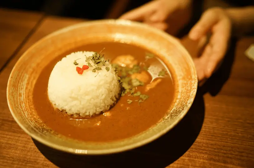 A cozy, warm-toned close-up of a rustic bowl of Japanese curry rice, featuring a neat mound of white rice garnished with herbs sitting in a generous pool of rich brown curry.