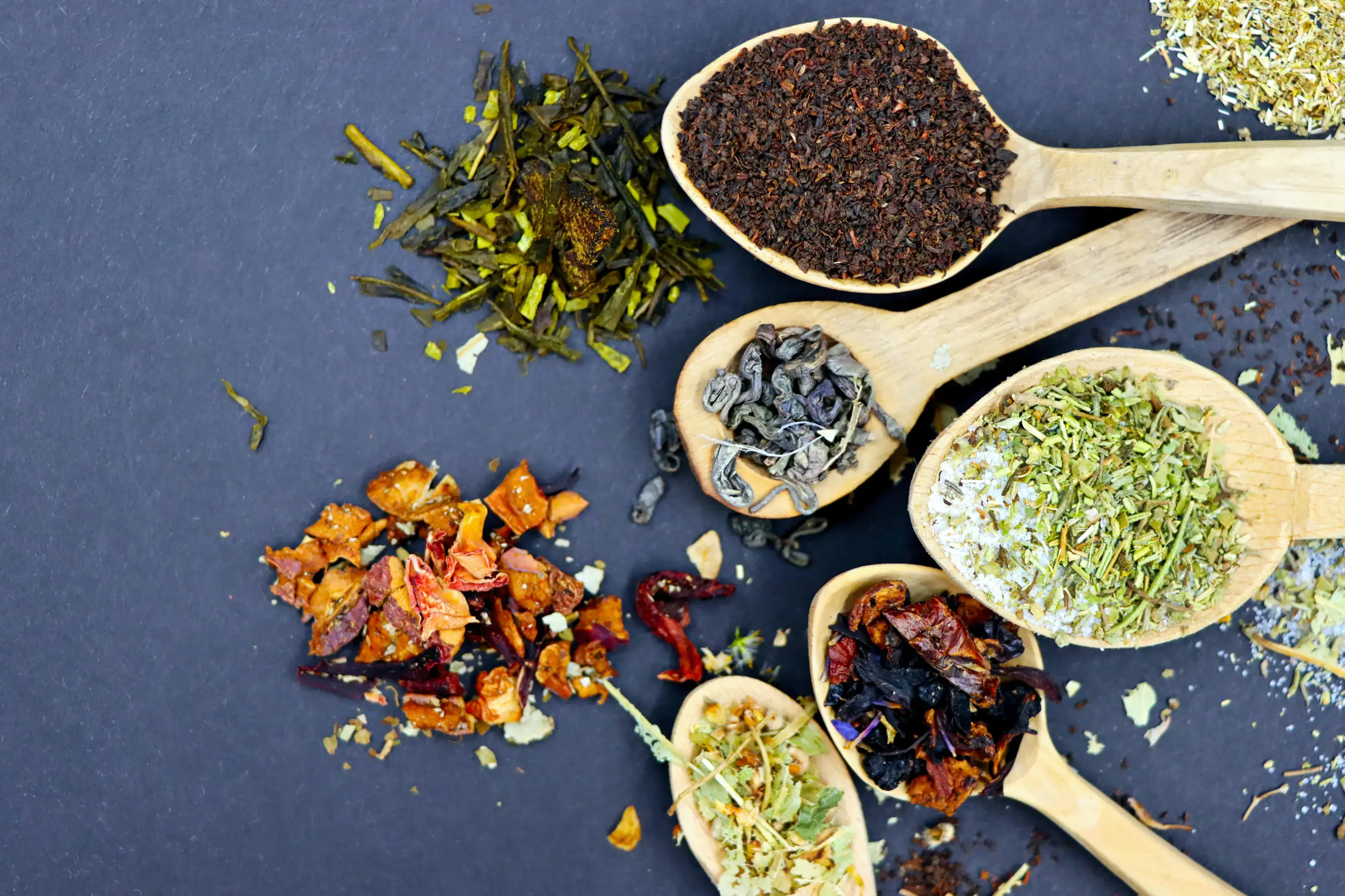 An overhead view showcases an assortment of loose-leaf teas and dried herbs arranged in wooden spoons and loose piles against a dark gray background. The varieties include green tea leaves, finely ground black tea, rolled leaves, and colorful fruit and floral blends.
