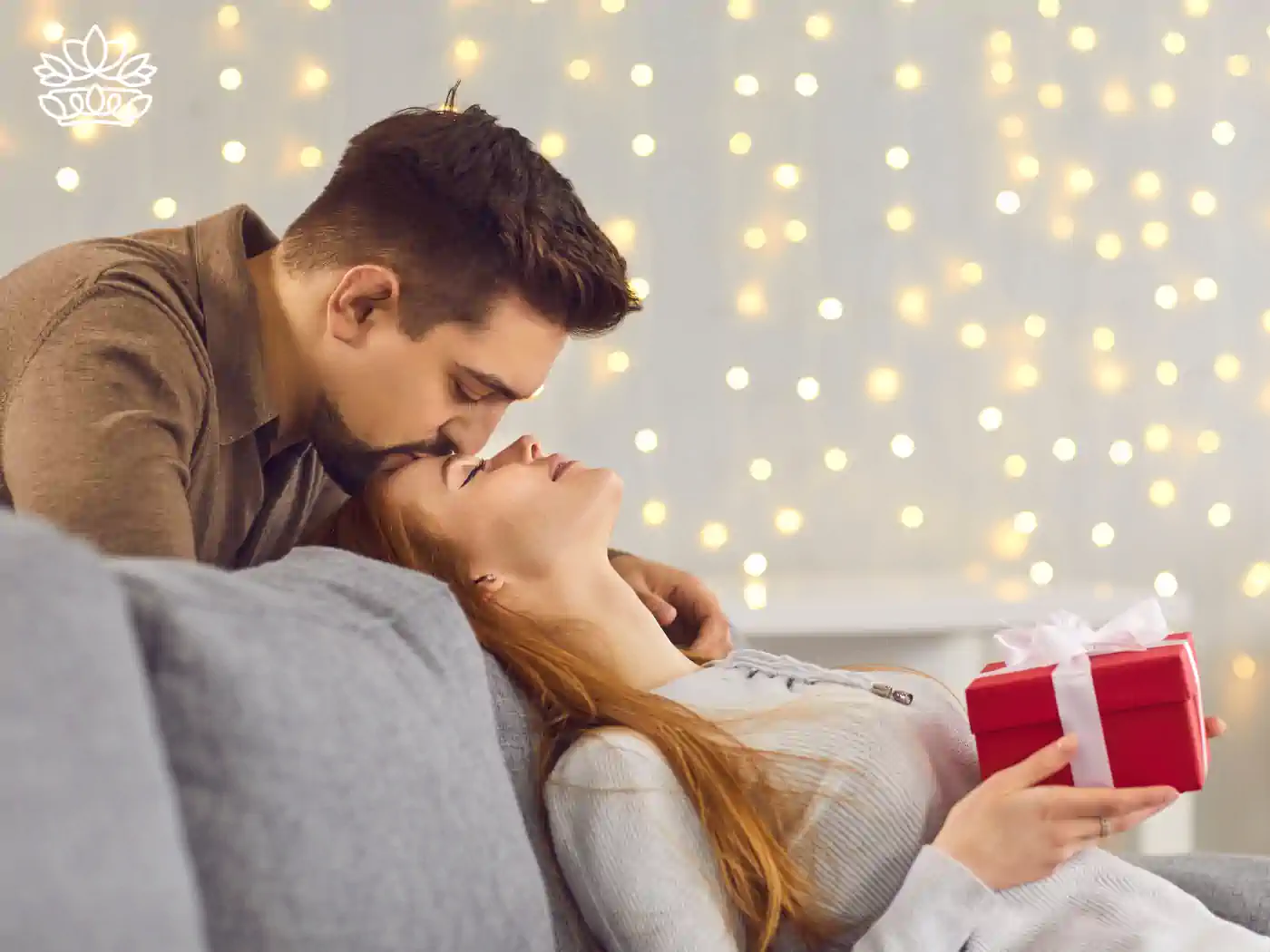Romantic couple sharing a tender moment with a Valentine's Day gift, surrounded by festive lights. Fabulous Flowers and Gifts.