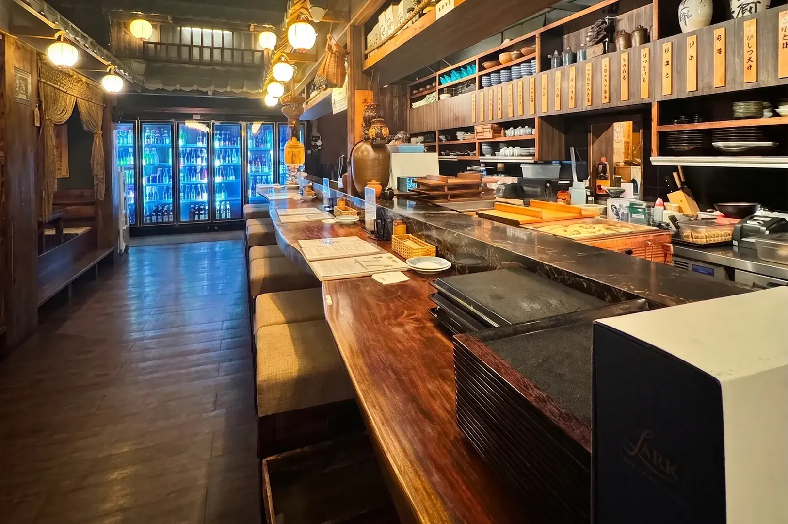 Interior of a cozy Japanese izakaya featuring a long wooden bar counter, traditional lighting, and shelves stocked with drinks.