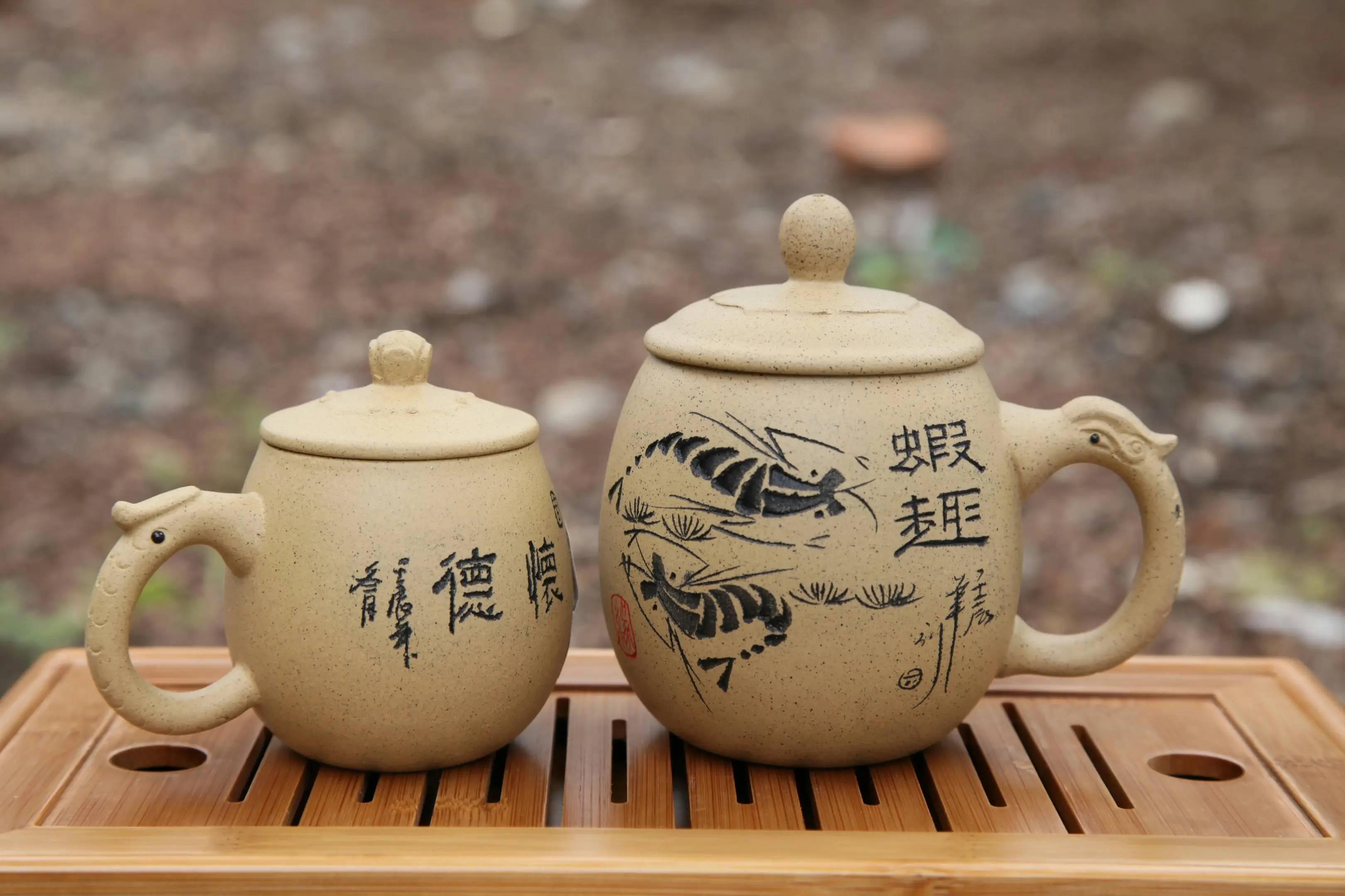 Two lidded ceramic mugs with ornate handles sit side-by-side on a slatted wooden tea tray. Both vessels feature intricate calligraphy and ink-style illustrations of shrimp, set against a blurred outdoor background.
