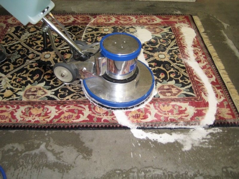 A rotary carpet cleaning machine with a chrome body and blue trim is applying foam onto an ornate red, black, and cream Persian rug. The rug is laid on a concrete floor with visible wet patches around it.
