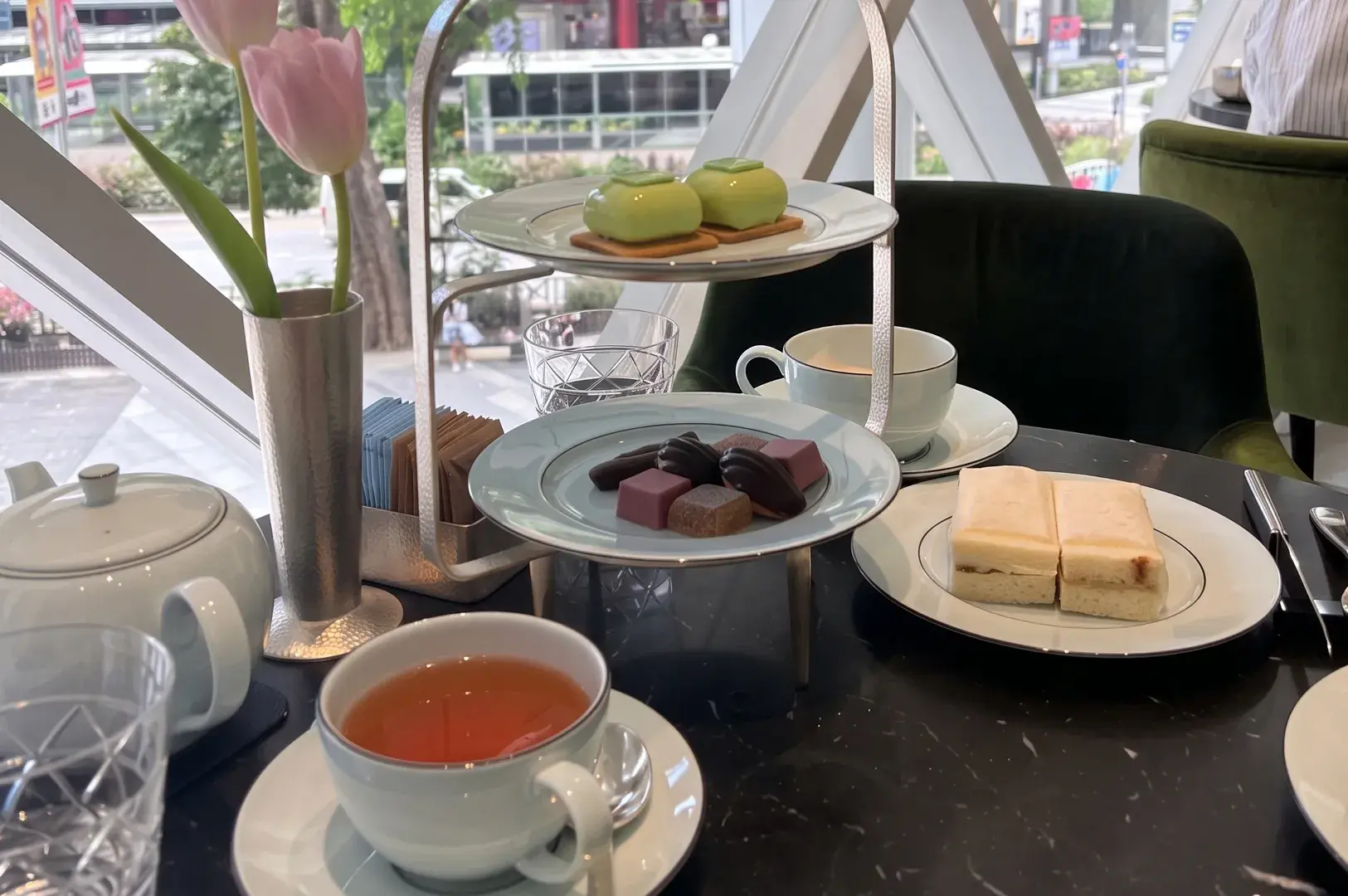Short Alt Text (Best for general web/social media) Afternoon tea setup with a silver tier of green pastries and chocolates, tea cups, sandwiches, and a vase with pink tulips by a window.
