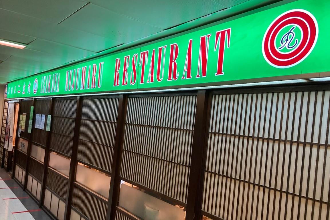 The image shows the exterior of Izakaya Nijumaru Restaurant, featuring a long, vibrant green sign with red and white lettering. The storefront is designed with traditional Japanese-style sliding wooden lattice doors and frosted glass panels.