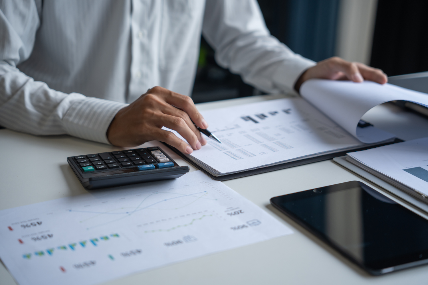 A professional reviews financial documents at a desk, using a calculator and tablet to analyze data and perform a professional business valuation.