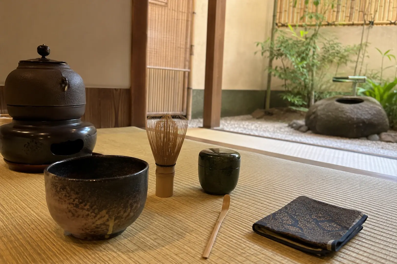 A low-angle, detailed shot of tea utensils meticulously arranged on a tatami mat floor. In the immediate foreground, a dark, textured ceramic tea bowl sits next to a standing bamboo whisk, a small dark green lacquer tea caddy, a slender bamboo tea scoop, and a neatly folded patterned cloth. To the left, a heavy iron kettle rests on a dark brazier. The background opens up to a tranquil, gravel-lined courtyard garden featuring a large, rounded stone water basin and lush green bamboo stalks behind a traditional wooden partition.