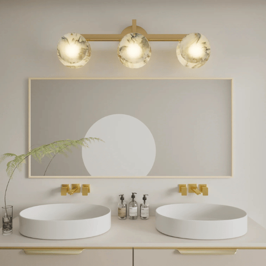 Modern bathroom vanity with two round white vessel sinks and gold wall-mounted faucets. Above, a large rectangular mirror is framed in light wood, topped with a three-light fixture featuring frosted glass globes with marbled patterns.