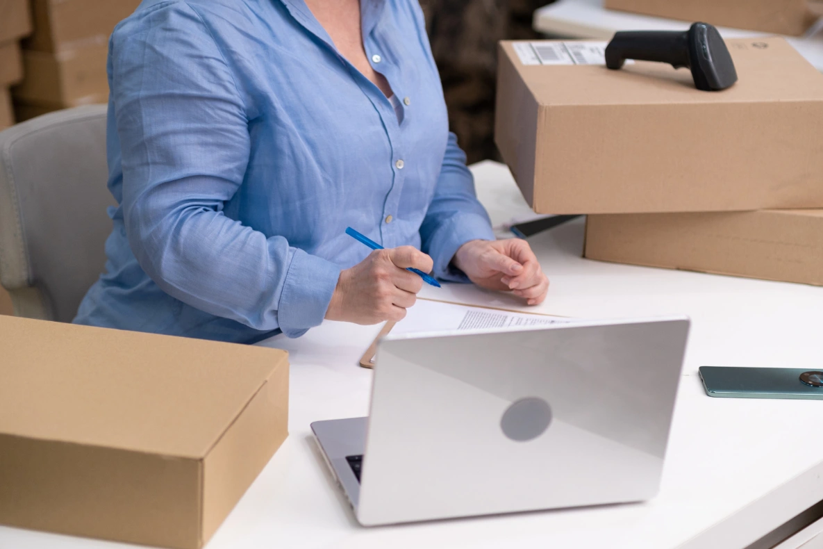 A person in a blue shirt is sitting at a desk with a laptop, pen, and paperwork. There are several cardboard boxes stacked around the desk, and a barcode scanner rests on one of the boxes.