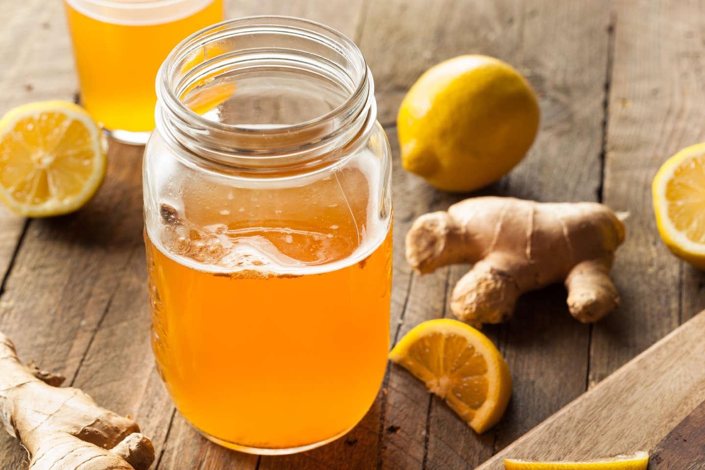 An open glass jar filled with an amber-colored liquid sits on a rustic wooden surface surrounded by fresh ginger roots and lemon slices. The bright, natural lighting highlights the vibrant colors of the ingredients, suggesting a fresh and healthy homemade beverage.