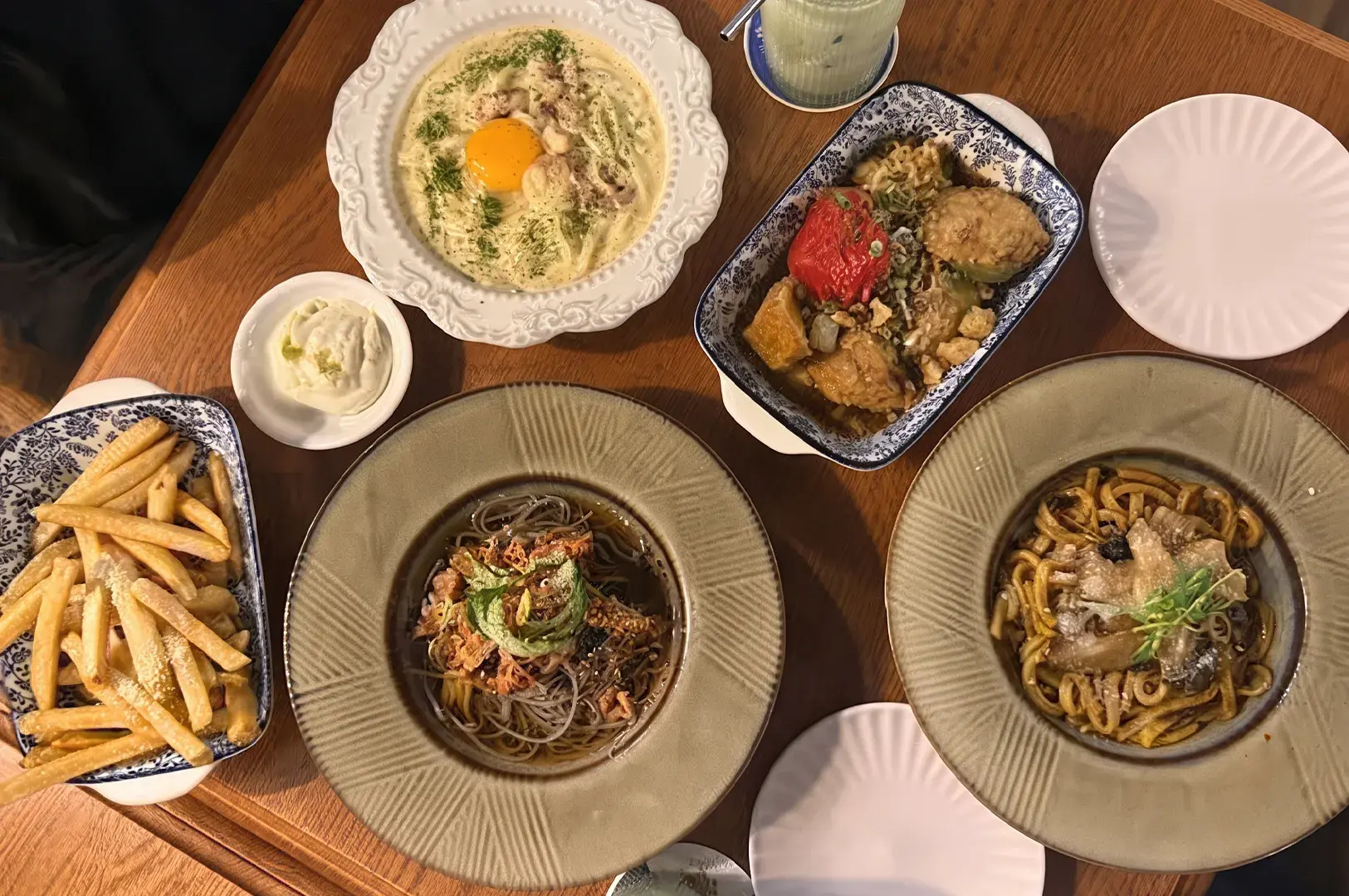 Overhead table view of a fusion meal featuring carbonara, soba, mushroom pasta, fries, and a casserole.