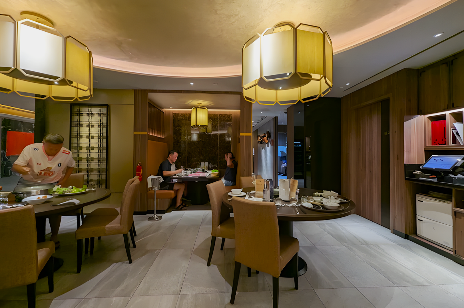 Upscale restaurant interior with modern lighting and wooden decor. People dine at round tables, creating a cozy, elegant atmosphere. Plates and tableware are neatly arranged.