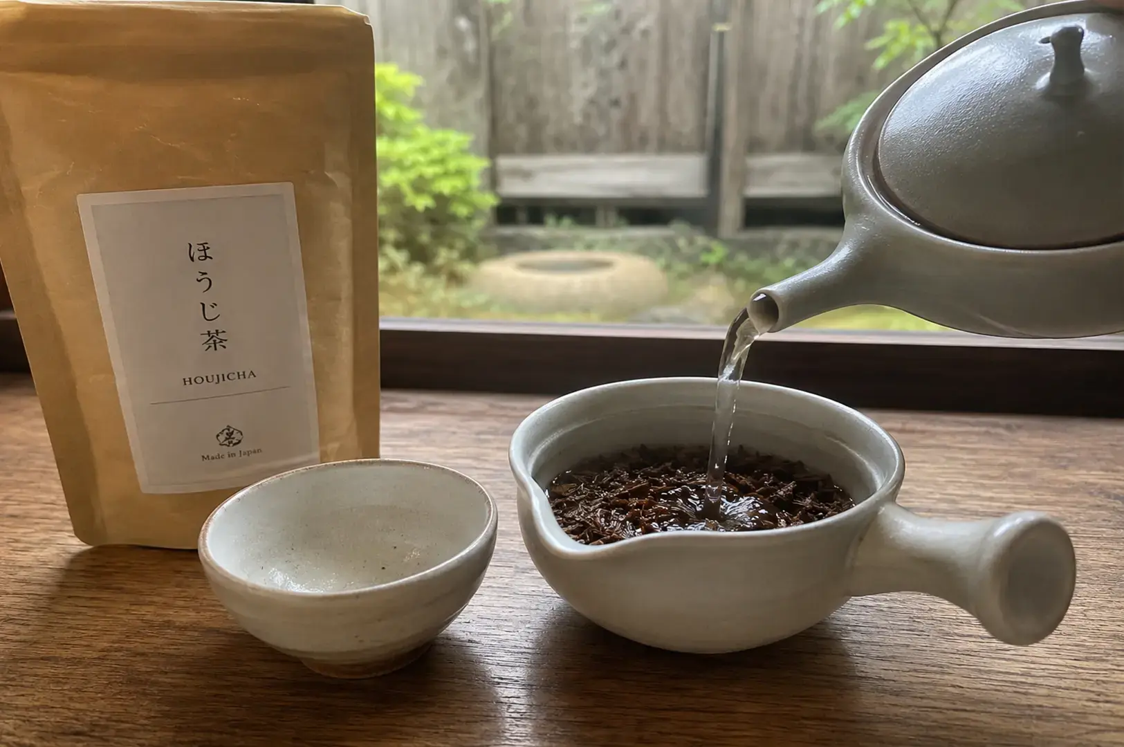 A light-colored ceramic teapot pours steaming water into a side-handled tea bowl filled with loose-leaf Hojicha, positioned next to a matching empty cup and a paper bag of "Hojicha" tea with a Japanese garden visible in the blurred background.