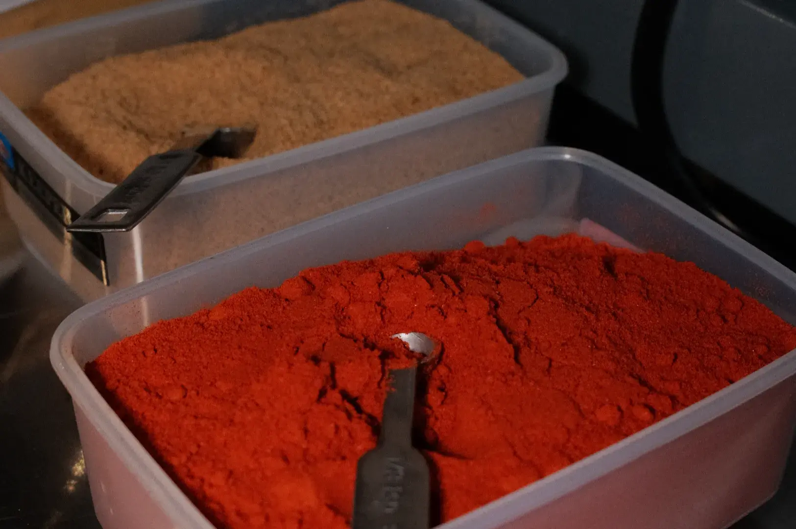Two clear plastic bins of ground spices: vivid red chili powder in front and tan-brown spice behind, each with a scoop, suggesting a market or kitchen display.