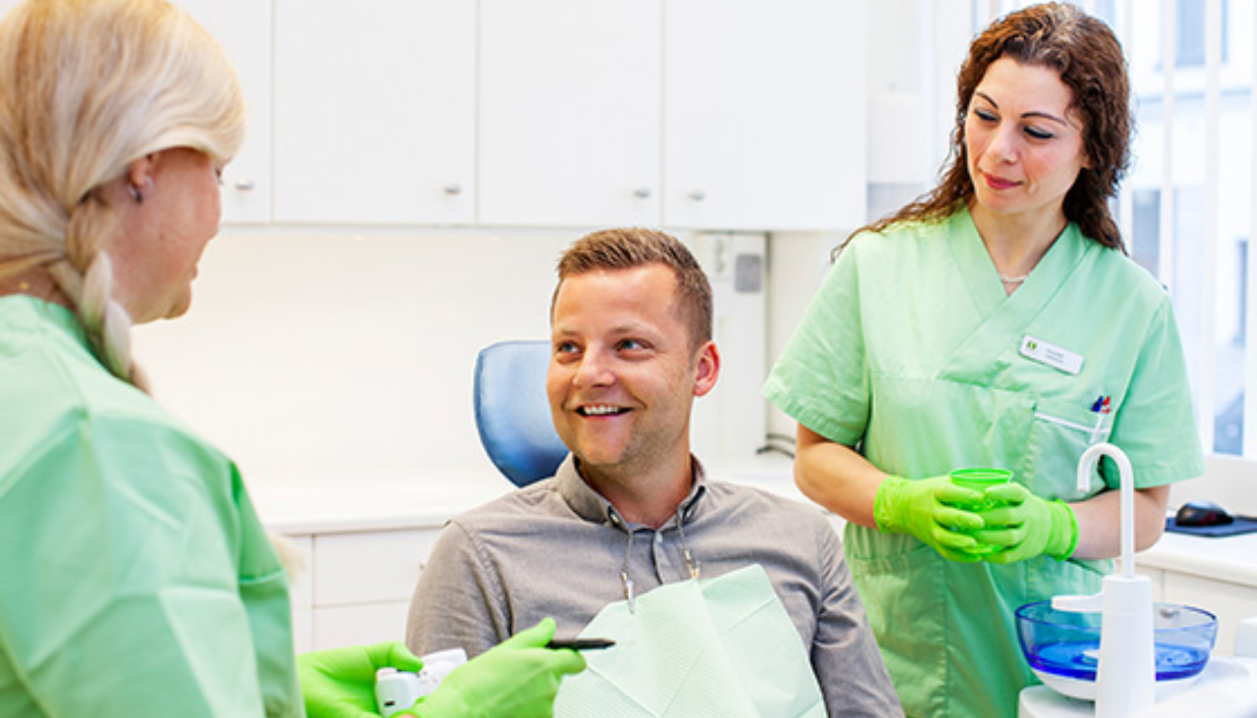 Mann sitter i tannlegestol og smiler mens to tannpleiere i grønn uniform snakker med ham.