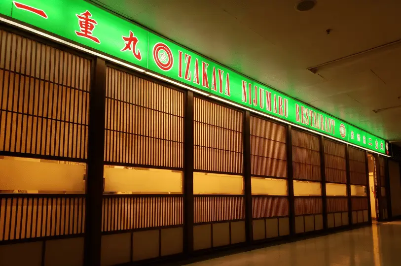 A long, glowing green neon sign for Izakaya Nijumaru Restaurant stretching across the traditional wooden shoji-style storefront in a Cuppage Plaza hallway.