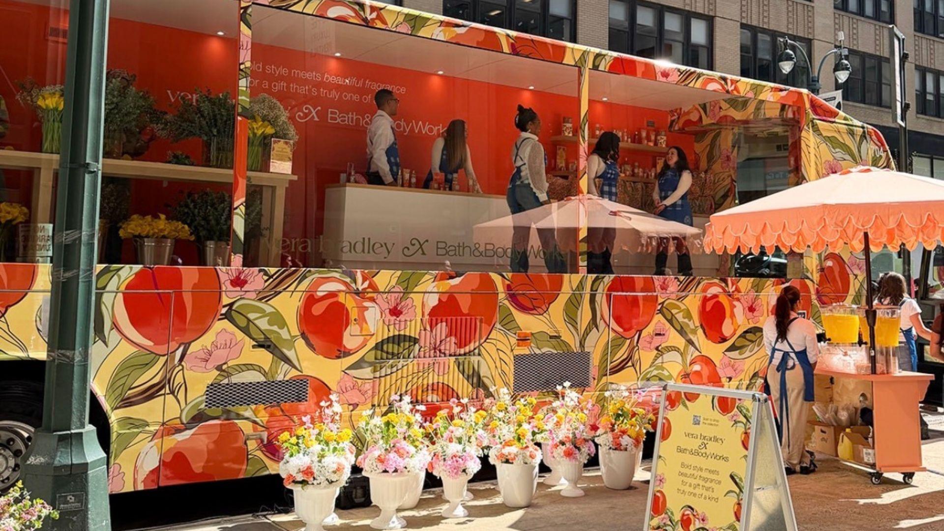 A big mobile bus that has a yellow background with peaches all over it, is parked on a busy street. Bath and Body Works workers. Bath and Body Works employees.