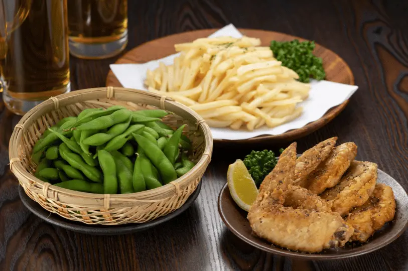 A trio of classic izakaya bar snacks—a woven basket of edamame, crispy french fries, and seasoned fried chicken wings—arranged next to tall glasses of beer on a dark table.