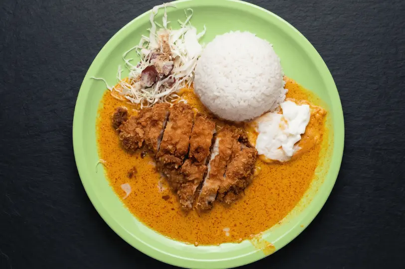 Top-down view of a bright green plate serving Japanese katsu curry on a dark slate background. The dish features a crispy, sliced fried pork cutlet resting in a pool of creamy orange curry sauce, accompanied by a mound of steamed white rice and a side of shredded cabbage salad.
