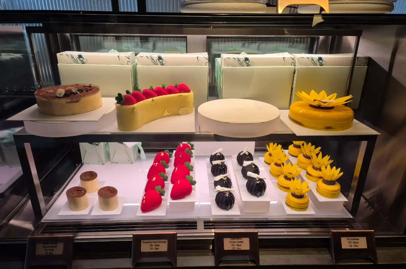 A glass display case showcases fine pastry creations organized on two lit shelves, including a long yellow mousse cake topped with red strawberries and gold leaf, a whole sunflower-shaped cake, and individual desserts resembling mini strawberries and dark glossy domes.