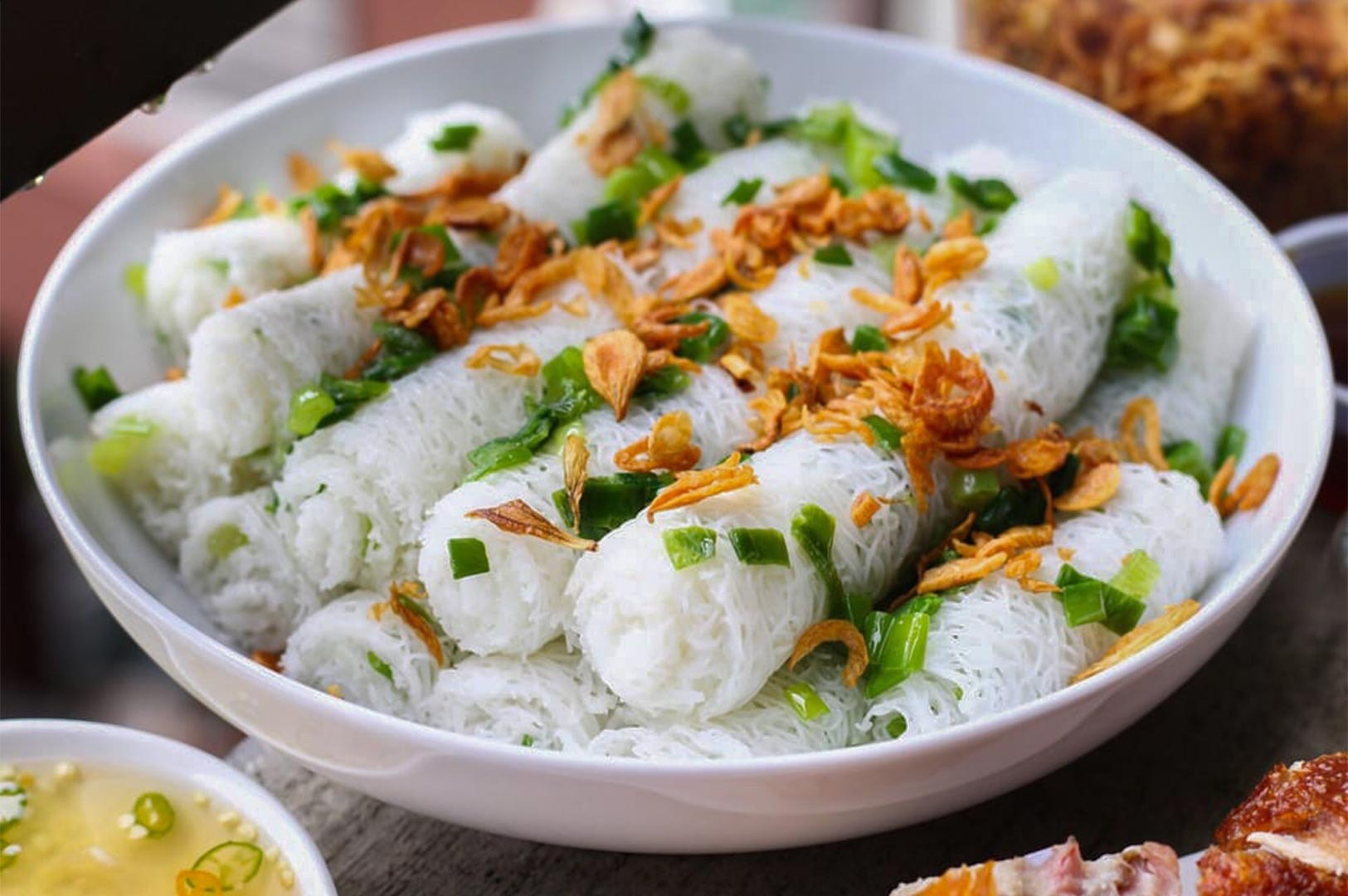 A white bowl filled with woven rice vermicelli bundles (Banh Hoi) topped with sautéed scallions and crispy fried shallots.