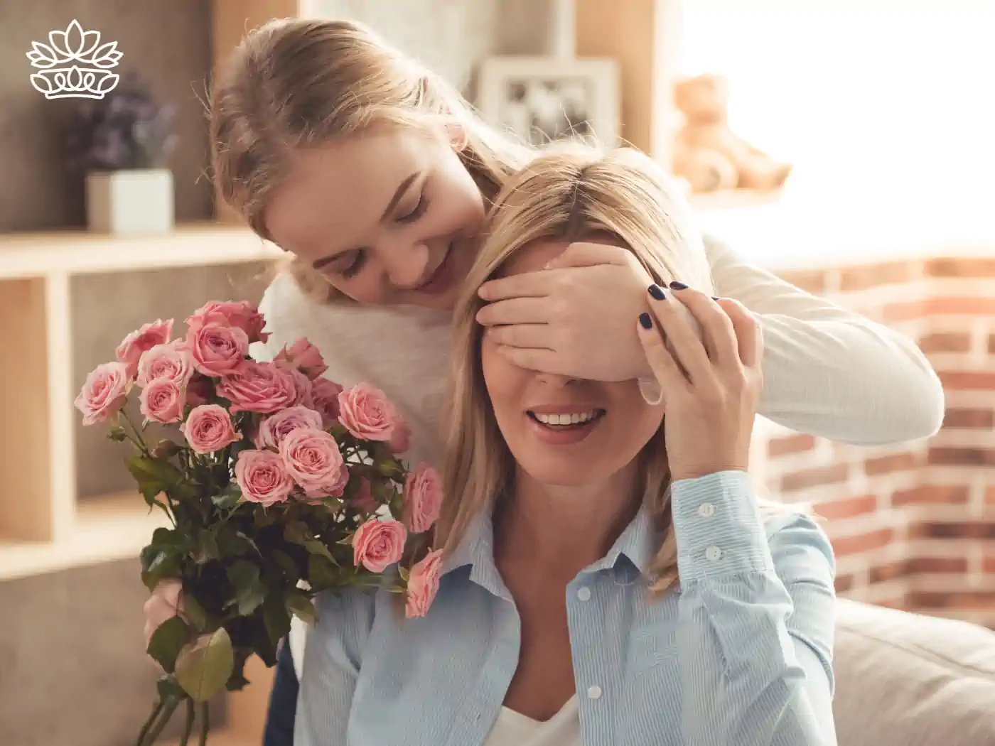 A young woman surprises another by covering her eyes from behind, holding a bouquet of pink roses, in a cozy, sunlit room. 