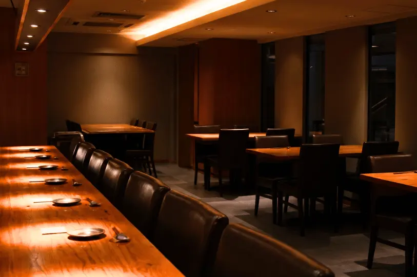 A dimly lit, upscale Japanese restaurant interior featuring a long polished wooden counter with leather chairs under warm spotlights, and darker group dining tables in the background.