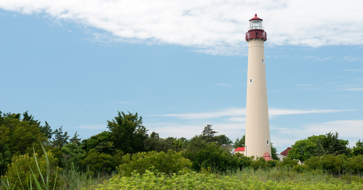 Cape May Lighthouse standing tall above lush green trees under a partly cloudy blue sky, located near North Cape May, New Jersey.