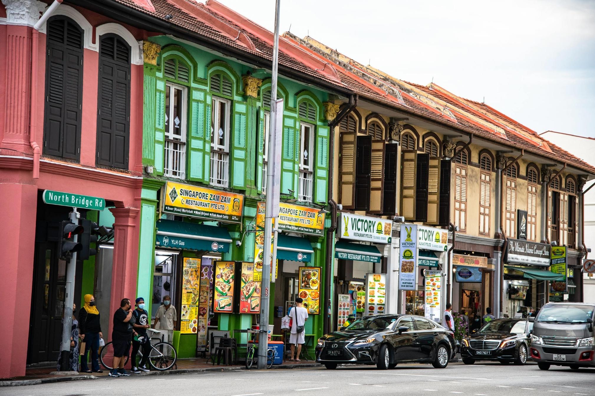 This street scene features a row of colorful shophouses, highlighted by the bright green facade of the "Singapore Zam Zam Restaurant" and the adjacent "Victory Restaurant" located on North Bridge Road. Pedestrians and a cyclist wait at the street corner while cars drive past these historic dining establishments.