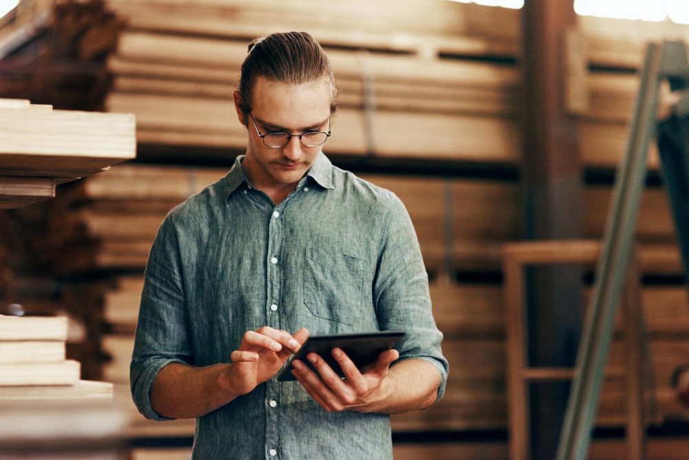 A carpenter standing in front of wood in workshop for manufacturing using internet protocol on a mobile device. 