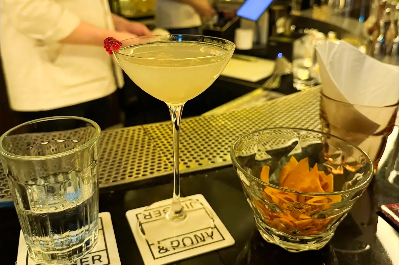 Close-up eye-level shot of a pale yellow cocktail served in a stemmed glass on a bar counter, with water glass, snack bowl, bar mats, and a bartender station visible in the background.