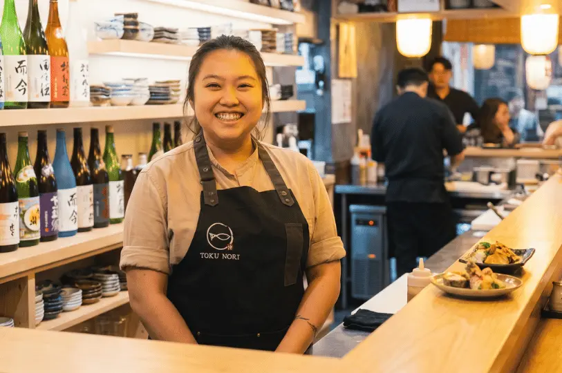 A smiling female chef wearing a black Toku Nori apron stands proudly behind a light wood sushi counter, with neatly arranged sake bottles on illuminated shelves behind her.