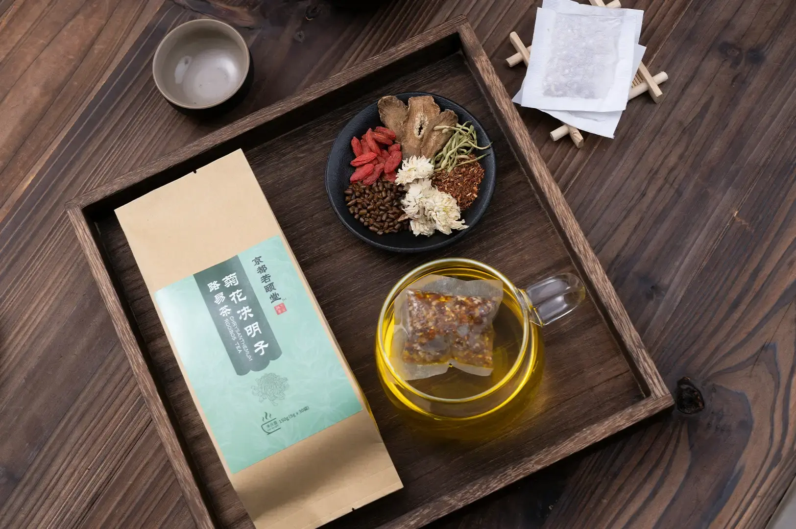An overhead shot displays a dark wooden tray containing a craft paper tea package labeled Chrysanthemum Rooibos Tea, a clear glass cup with steeping golden-yellow tea, and a small black dish featuring raw ingredients like goji berries, cassia seeds, and dried chrysanthemum blossoms. An empty ceramic teacup and tea filter bags sit on the surrounding dark wood background.