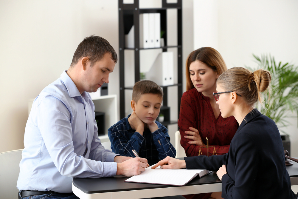 A dad, mom and son sit at a table with a lawyer, signing documents