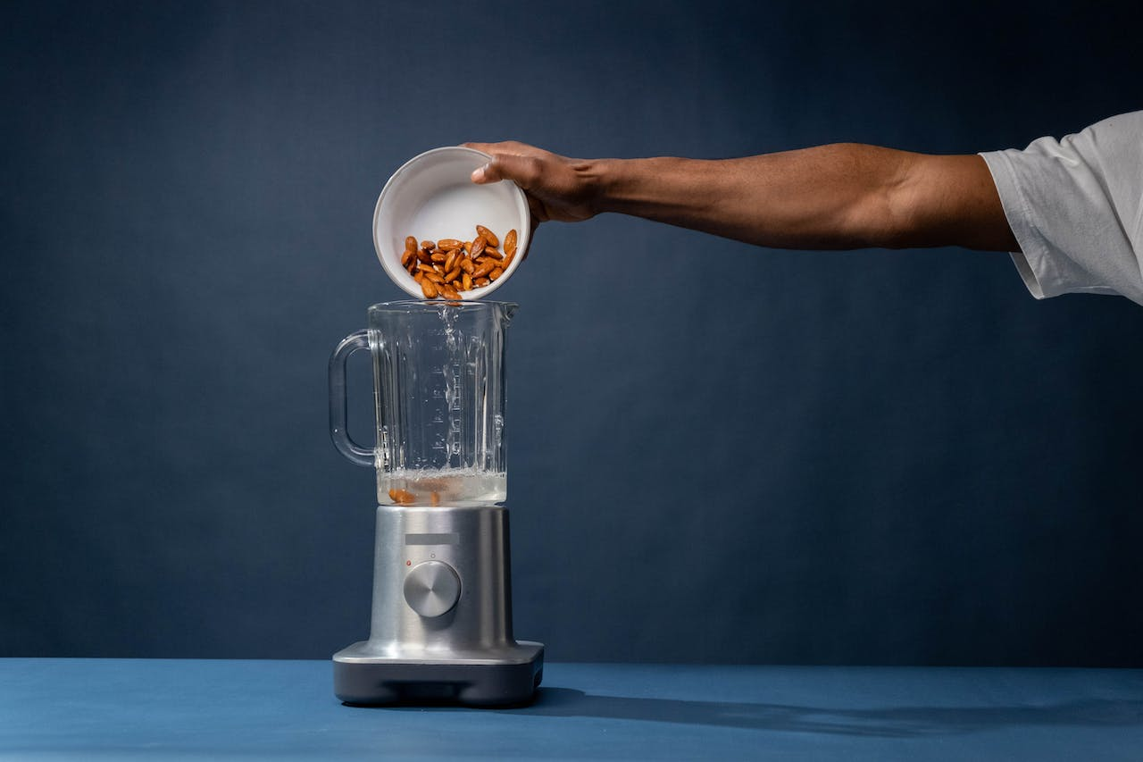 Uma mão está despejando algo que parece ser amendoins em um liquidificador vazio, indicando o passo inicial de uma receita que pode incluir esses ingredientes.