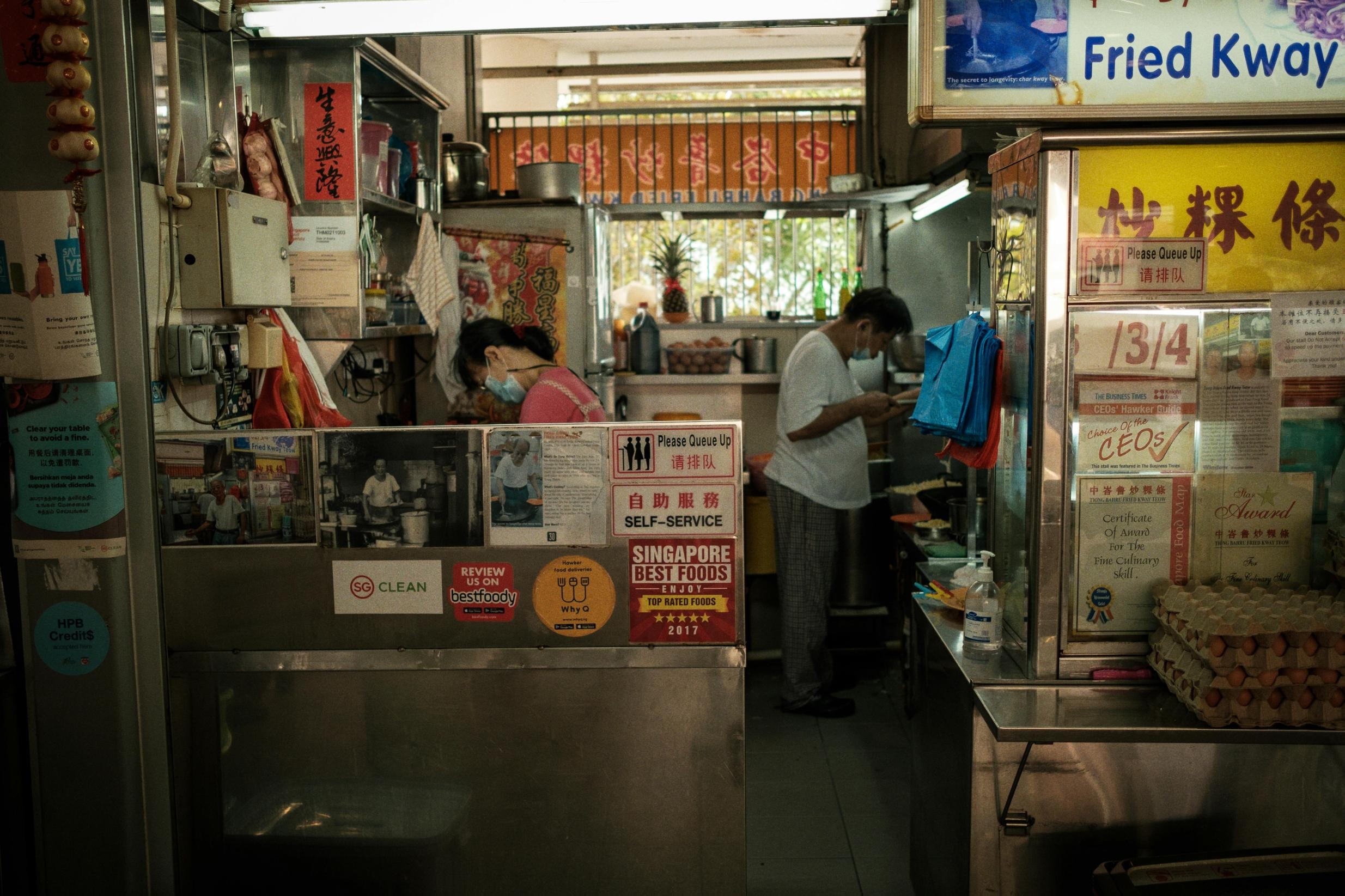  In a cramped but organized food stall, two workers wearing face masks prepare meals behind a counter adorned with "Singapore Best Foods" awards and "Self-Service" signs. The workspace is filled with culinary essentials, including stacks of egg cartons, fresh pineapples, and a menu board prominently featuring "Fried Kway Teow" in English and Chinese.