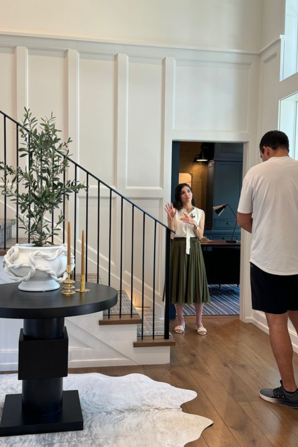 A bright foyer with crisp wall paneling, a black pedestal table styled with sculptural decor, and warm wood flooring beside an iron staircase railing.