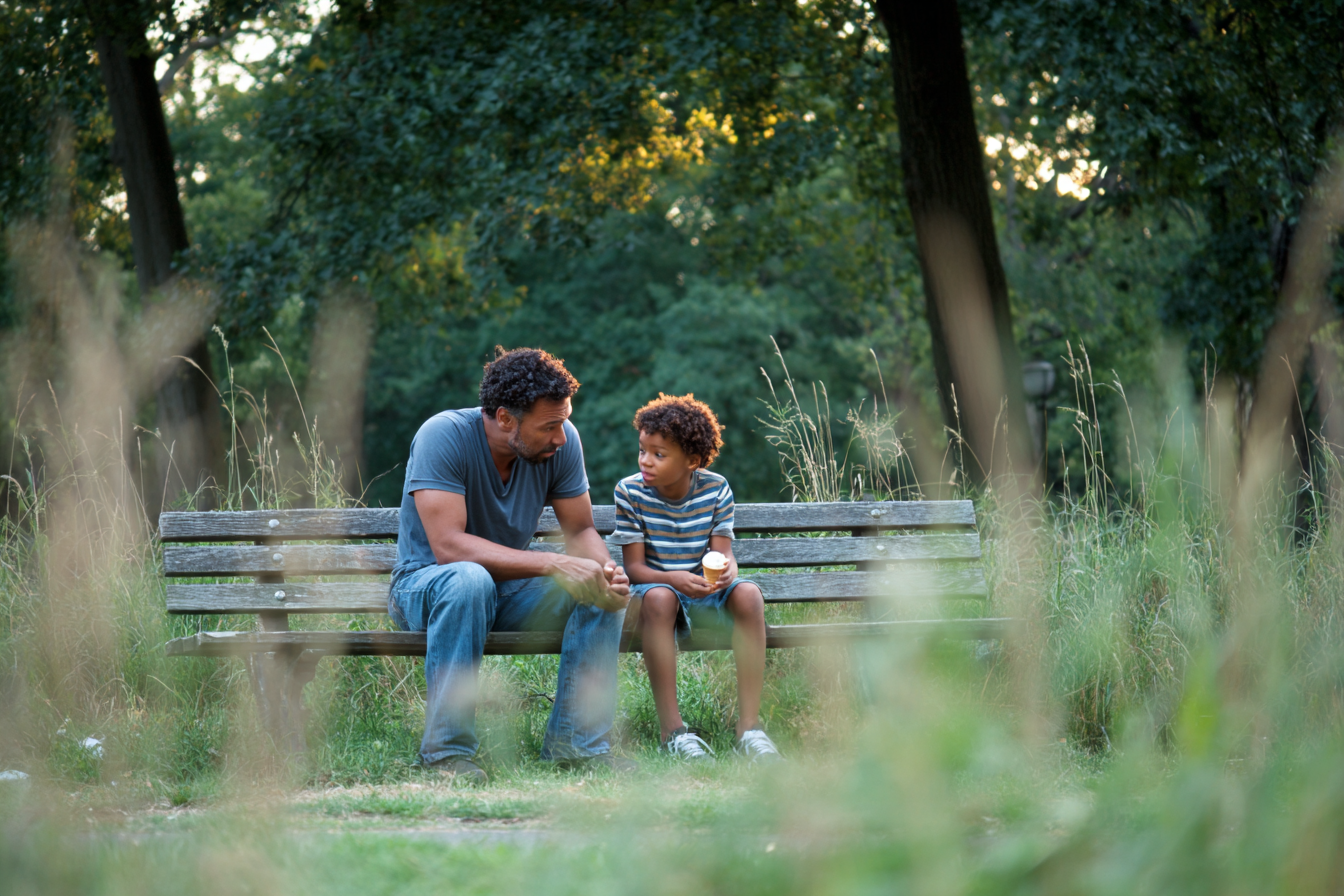 A father and his young son sitting together on a Prospect Park bench in early summer afternoon light