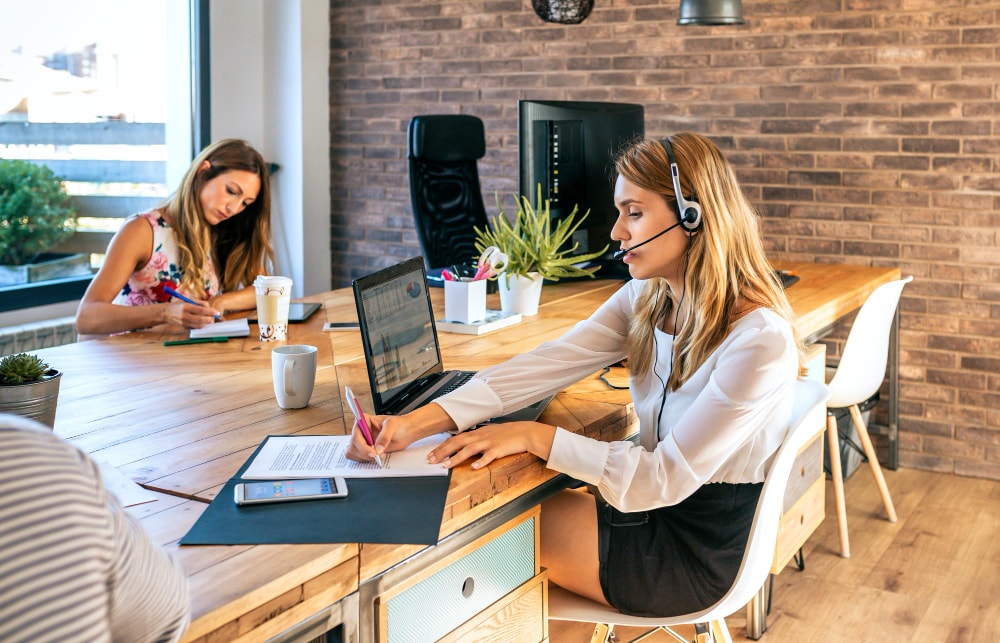 A woman in a headset takes notes while working at a long wooden desk with a laptop and plant decor; another woman works in the background.